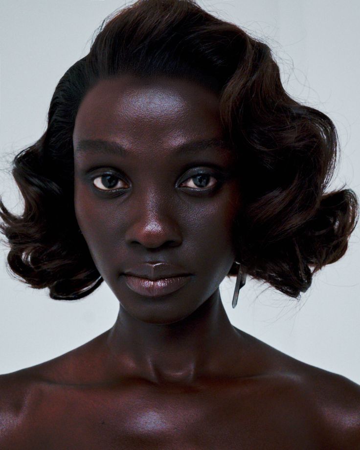 the image is a closeup portrait of a black woman with dark skin. she has a styled, short, wavy hairstyle reminiscent of the 1920s.  her expression is serious and composed. the lighting is soft and highlights her features, particularly her skin which has a subtle, healthy glow.  she is wearing simple earrings and is looking directly at the camera. the background is plain and uncluttered, drawing focus entirely to the subject. the overall feel is one of elegance and sophisticated beauty.