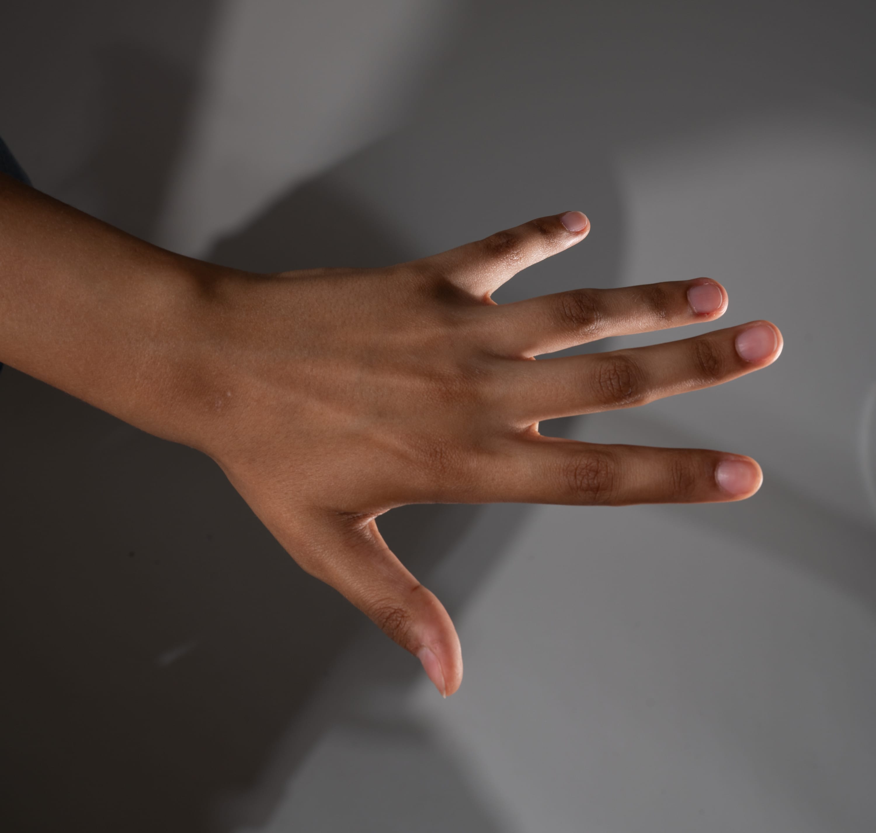 a closeup view of a hand, partially illuminated and casting a distinct shadow on a neutralcolored background. the hand, belonging to a person of color, is positioned with fingers extended, showcasing its natural curvature and skin tone.  the lighting accentuates the texture and details of the hand, highlighting the subtle variations in color and the play of light and shadow.