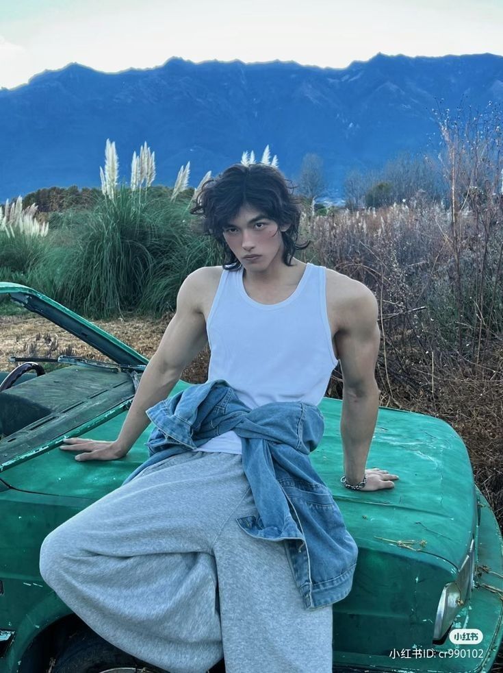 a young asian man is sitting on the hood of a green car. he's wearing a white tank top, grey sweatpants, and a blue denim jacket. the backdrop is a mountain range with a blue sky and green grass fields.