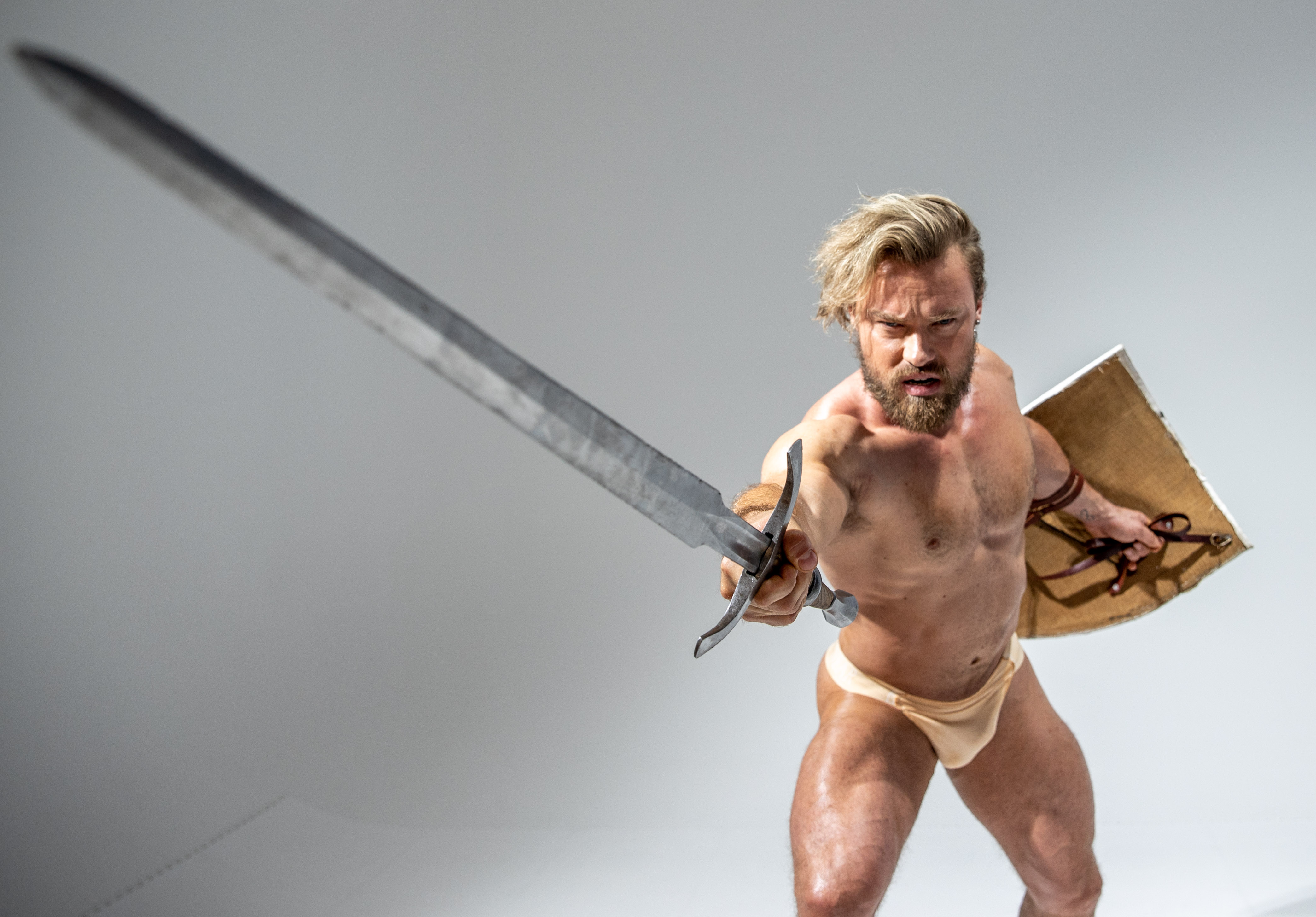 a muscular man with a beard and long blond hair, clad only in beige underwear, fiercely brandishes a large, gleaming sword in a fighting stance. he grips the sword with both hands, its point directed menacingly towards the viewer.  he also holds a round shield made of wood and leather. the backdrop is a solid, bright gray, emphasizing the dramatic pose and the intensity of the warrior.  the low camera angle further accentuates his power and dominance.