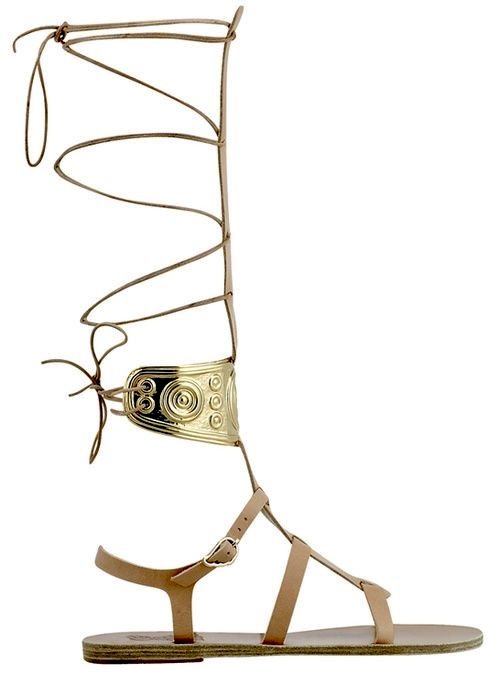 a single, highresolution image showcases a tancolored leather gladiator sandal. the sandal features a flat sole with multiple thin straps across the foot, a decorative gold cuff detail with circular patterns wrapping around the ankle, and a laceup design extending high up the leg. the style is a women's fashion item, suitable for warm weather.