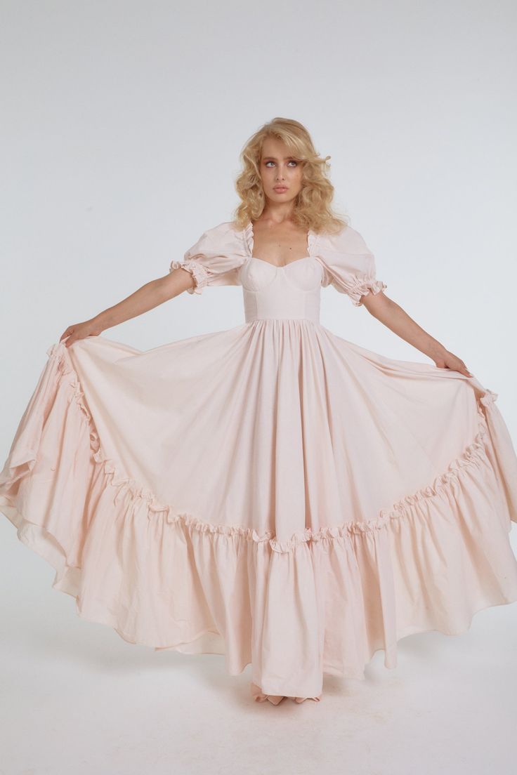 a young blonde woman poses in a flowing, romantic, pale pink dress. the dress features a sweetheart neckline, puff sleeves, and a tiered, ruffled skirt. the background is a plain white studio backdrop. the lighting is soft and diffused, creating an ethereal and dreamy atmosphere.
