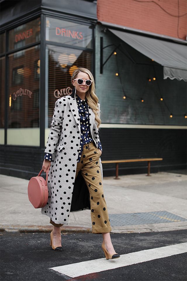 the image shows a stylish blonde woman walking down a city street. she's wearing a gray polka dot overcoat over a navy polka dot blouse and mustard yellow, polka dot cropped trousers. her accessories include round pink bag, beige twotone heels, and pinkframed sunglasses.  the background features a cafe or restaurant with a visible storefront. the overall style is casual chic, showcasing a coordinated and trendy autumn/fall outfit.