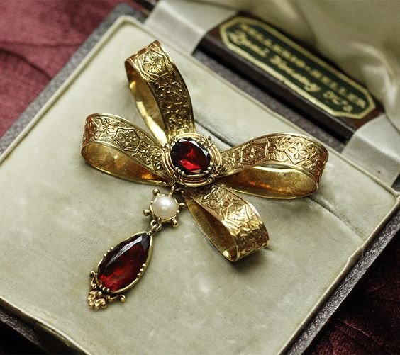 a gold bow brooch with intricate filigree details, adorned with two red garnets and a pearl drop, is displayed inside a velvetlined jewelry box. the box bears a label "audemar sellers  basel  dep. 023".