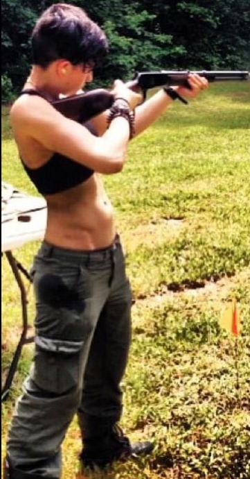 a woman is seen outdoors, aiming a rifle. she's wearing a bralette, green tactical pants, and boots. she is standing in a field, practicing target shooting. her short hair and bracelets are visible.