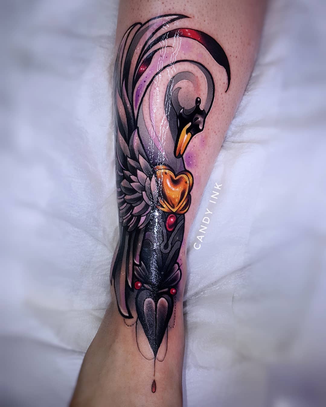 a neo traditional tattoo of a swan with a heartshaped locket on its chest. the swan is black and grey with red and gold accents. the tattoo is on the back of someone's lower leg, just above the ankle.