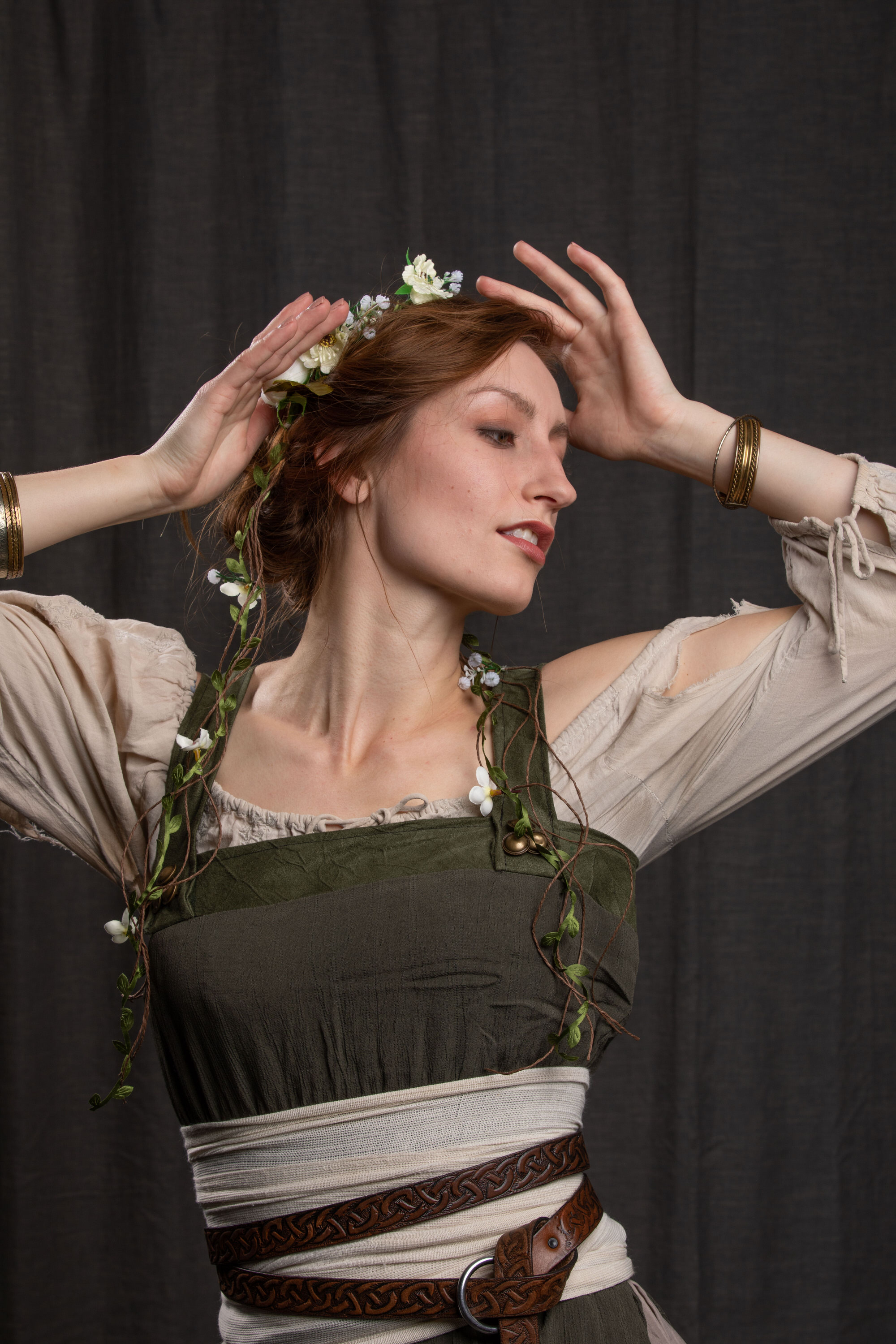 a woman with auburn hair styled in an updo with a flower crown poses against a dark gray backdrop. she wears a green, white, and brown medievalstyle costume, accented with leather belts and floral embellishments. her expression is serene as she looks off to the side, her hands raised to her head. the artificial lighting casts soft shadows and highlights on her features and attire.
