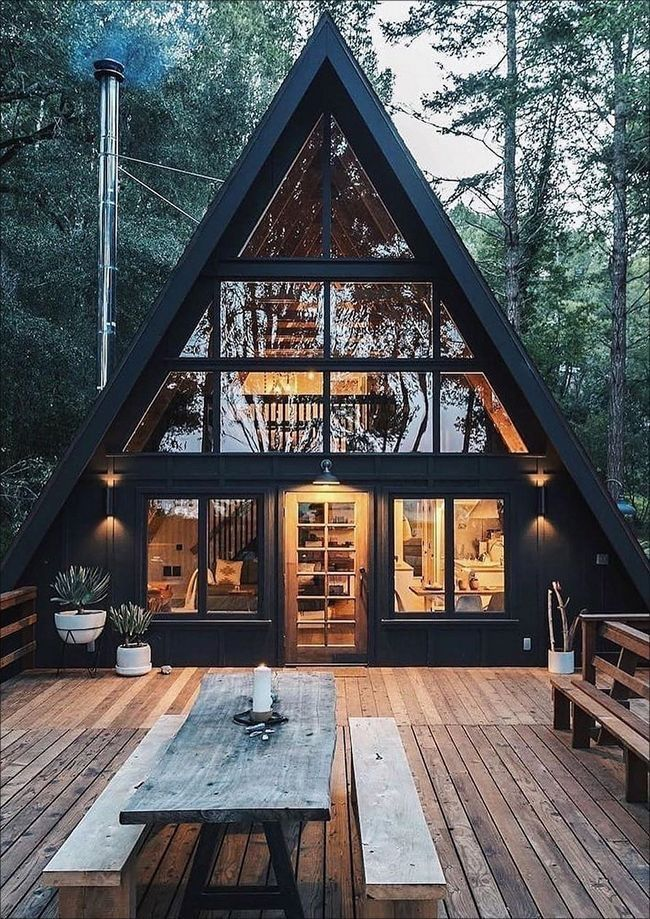 the image showcases a stunning aframe cabin nestled in a dense forest. the cabin's exterior is painted a sleek black, contrasting beautifully with the surrounding greenery.  large, expansive windows dominate the front of the cabin, allowing ample natural light to illuminate the interior, which is visible through the glass.  the cabin features a spacious wooden deck with a rusticstyle picnic table and benches providing comfortable outdoor seating. potted plants add to the cabin's inviting atmosphere. the overall scene conveys a sense of tranquility, comfort, and seclusion, ideal for a peaceful getaway. the time appears to be evening or night, as the interior lights are on and cast a warm glow.