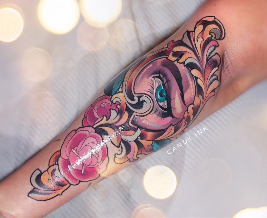 a forearm tattoo of an eye surrounded by decorative gold filigree and pink flowers. the tattoo is done in a neotraditional style with bold lines and bright colors. the tattoo is on a person's left forearm. the photo is taken from above looking down at the tattoo. the background is blurry and out of focus.