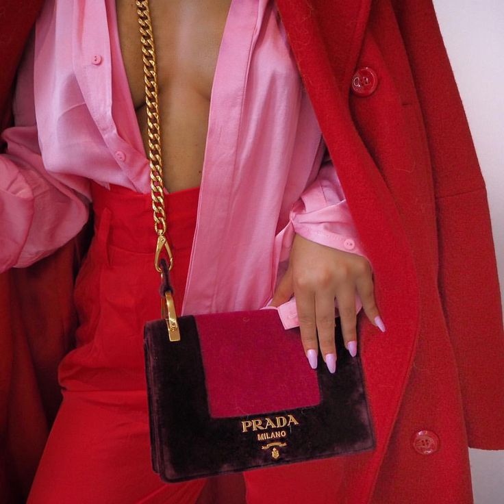 closeup view of a woman's stylish outfit. she is wearing a red wool coat over a pink silk shirt and matching red pants.  the focal point is a prada velvet handbag in shades of burgundy and dark brown with a gold chain strap. the woman's hands are visible, showcasing light purple nail polish. the overall aesthetic is luxurious and fashionable, emphasizing a colorcoordinated red and pink palette.