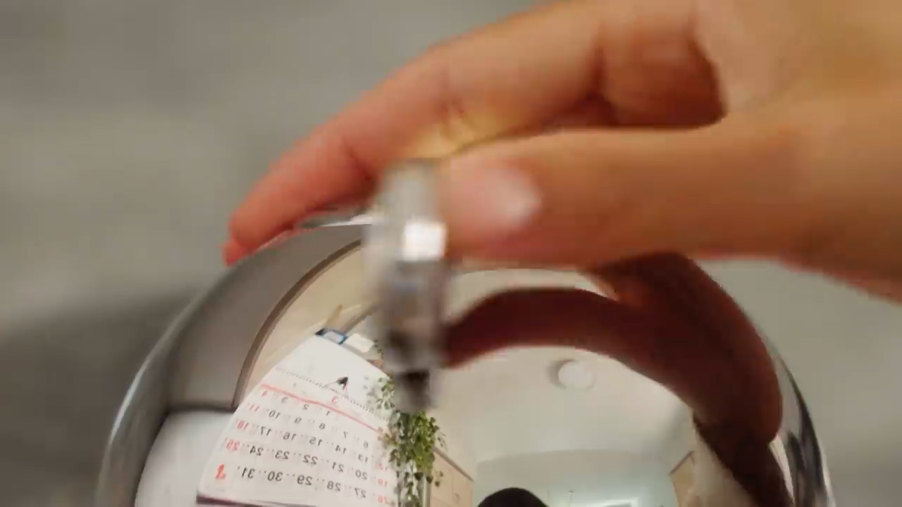 a hand wearing a ring is holding a metallic sphere. the reflection in the sphere shows a fisheye perspective of the room, including a calendar, a plant, and the person's arm.