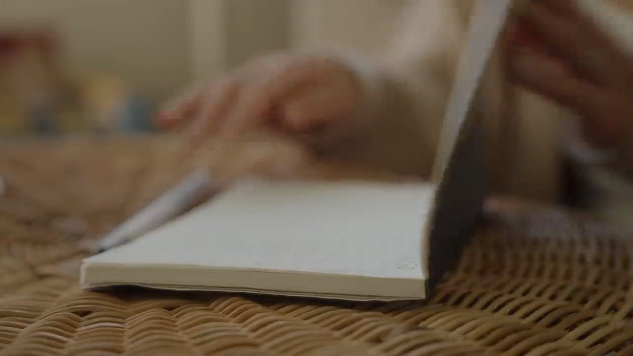 a closeup shot of a person's hands as they write in a notebook. the notebook is open and the pages are blank. the person is using a pen to write. the image is lit with natural light and has a soft, warm feel. the overall palette is neutral and calming, with shades of beige, brown, and cream.