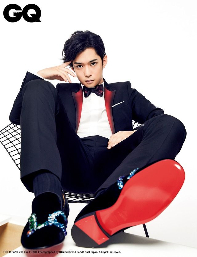 this is a photo of a man in a suit, sitting on a chair. he's wearing a black suit with a red and black tuxedo jacket. he's also wearing a bow tie and red sole shoes. the photo is taken from a low angle, looking up at the man.  the background is white and there's a gq magazine logo in the top left corner.