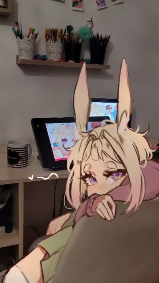 the image shows a digital drawing of a cute anime girl with bunny ears, seemingly overlaid onto a photograph of a reallife artist's workspace.  the workspace includes two drawing tablets, several containers filled with art pencils, and other art supplies neatly organized on a shelf and desk.  the anime girl is depicted in a cozy pose, seemingly hugging herself and looking slightly down. a mug sits on the desk beside the drawing tablets. the overall atmosphere is calm and creative.