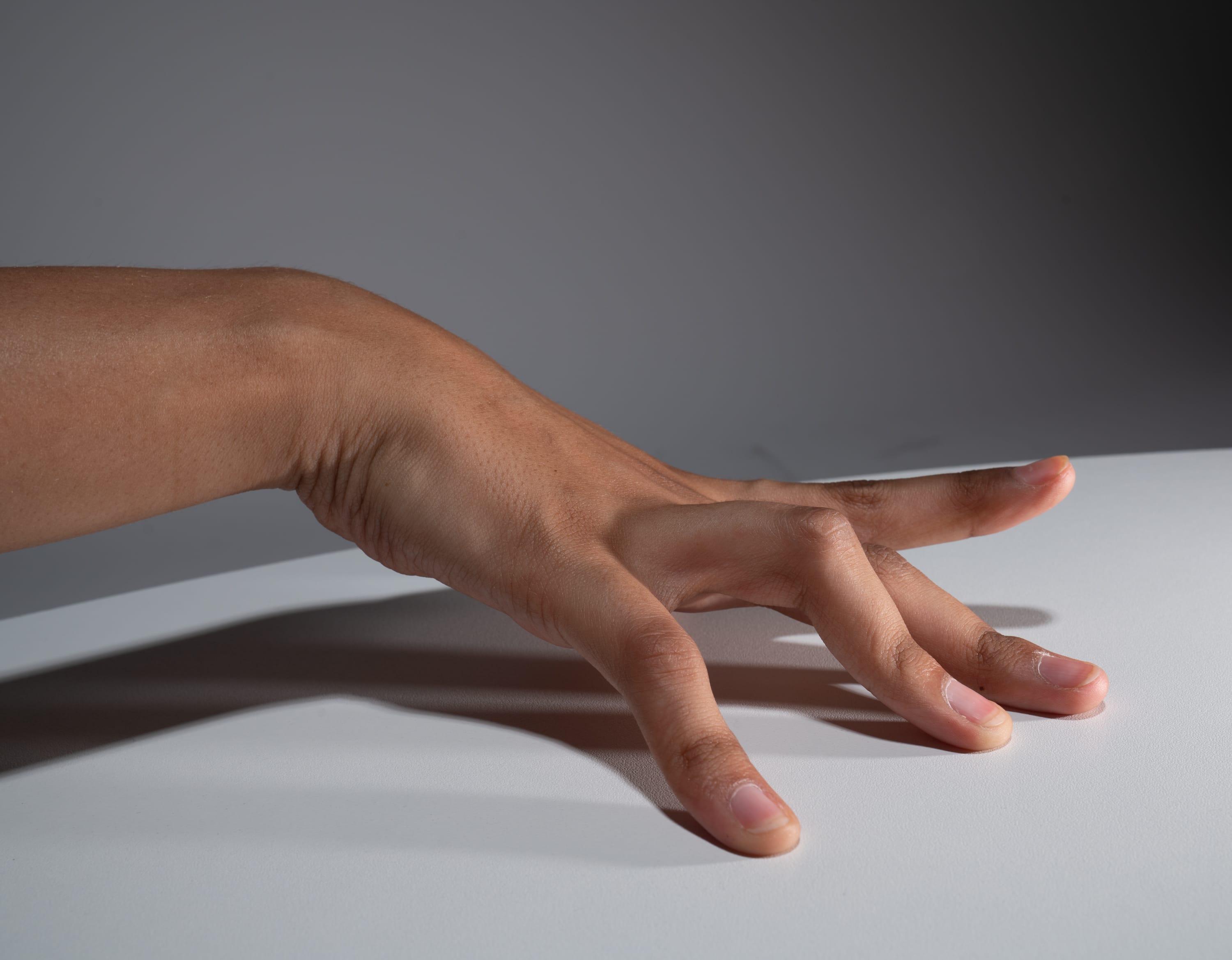 a hand with mediumdark skin tone is extended, fingers splayed, against a white surface. the background is a darker gray. the hand is lit from above.