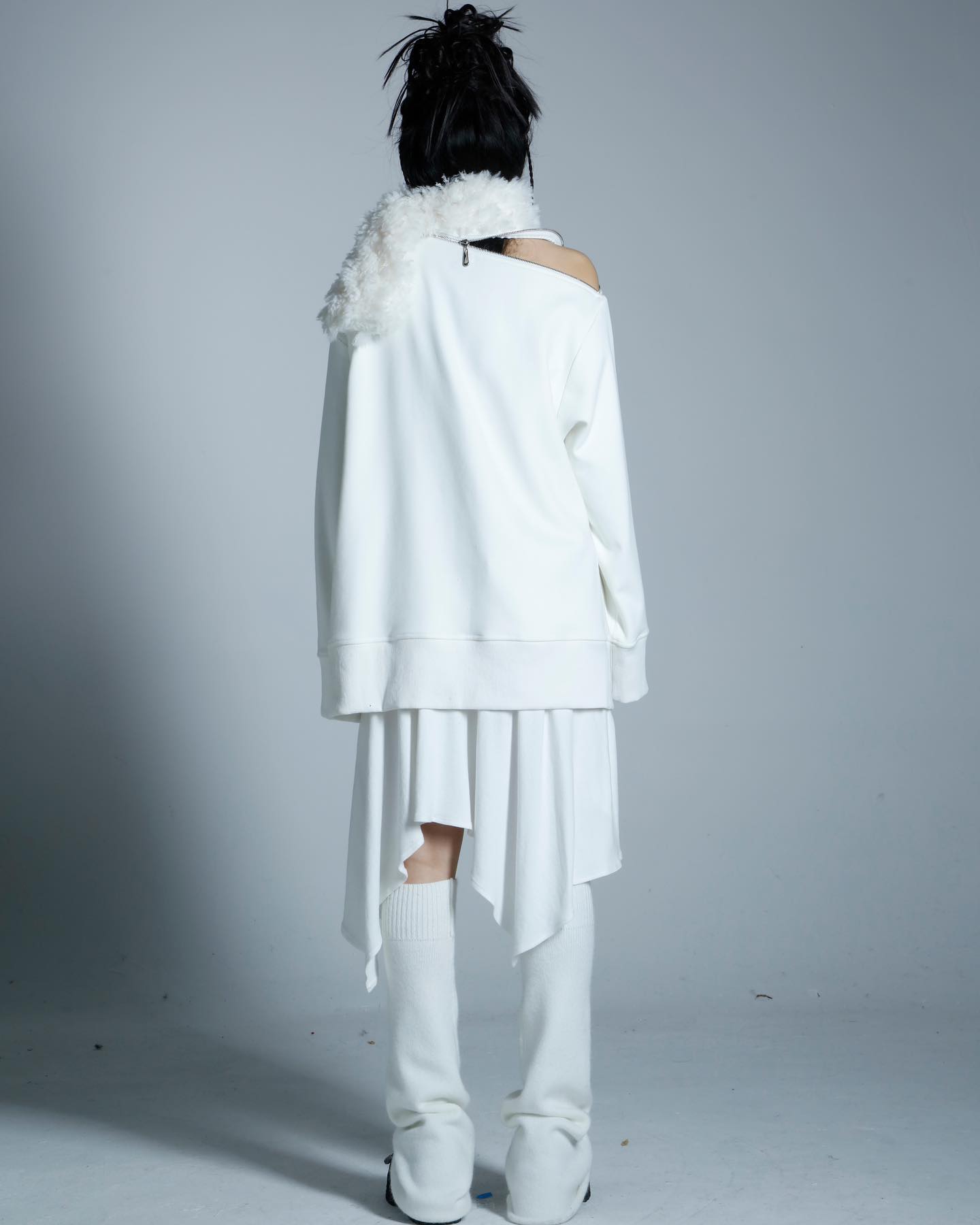 the image shows a rear view of a female model wearing an allwhite outfit. the outfit consists of an oversized, offtheshoulder sweatshirt layered over an asymmetrical skirt.  a fluffy white scarf is draped around the neck. the model is also wearing high white socks and white boots. the overall style is minimalist and contemporary, with a unique asymmetric design element in the skirt. the background is a plain, light grey, emphasizing the clothing. the photo appears to be a professional fashion shot.