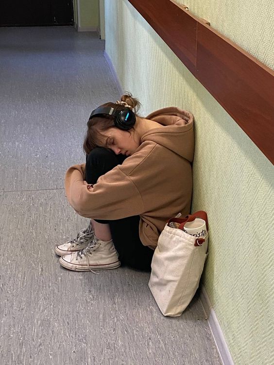 a young woman wearing a tan hoodie, black pants, and white sneakers sits huddled against a wall in a hallway. her head is down, resting on her knees, and her eyes are closed. she is wearing headphones and appears to be asleep. a white tote bag with brown straps rests on the floor beside her. the hallway is painted in muted green and brown tones, and the lighting suggests it is daytime.  the overall impression is of someone seeking a moment of rest or privacy in a busy environment.