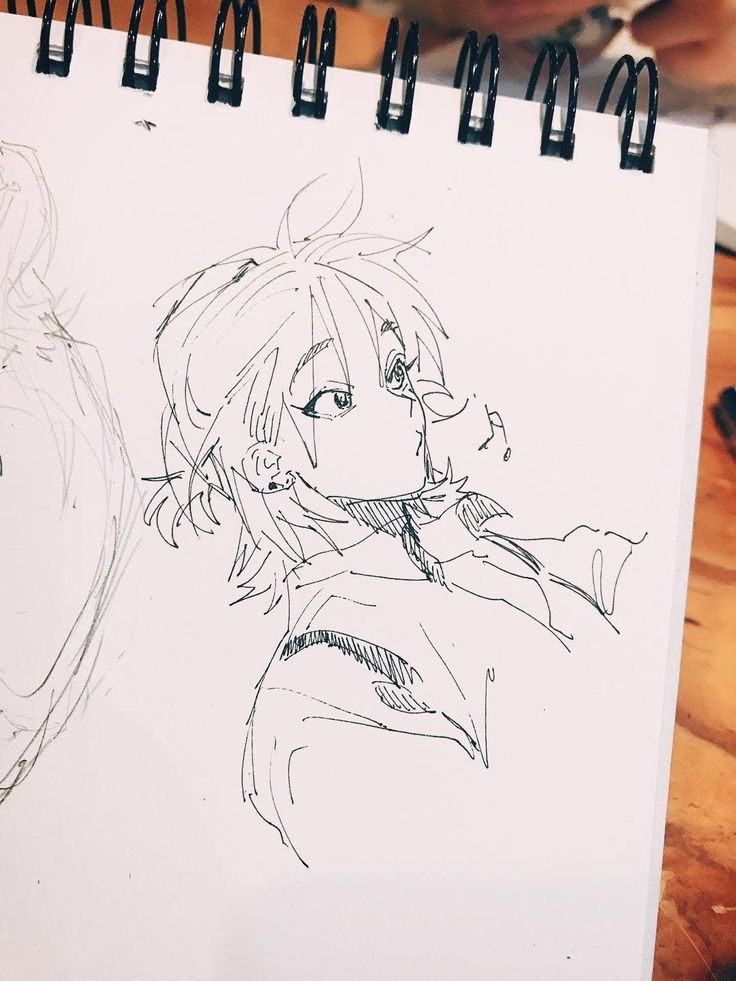 the image shows a closeup shot of a sketchbook page featuring a penandink sketch of an animestyle female character. the character is depicted from the shoulders up, with short, somewhat messy hair and a slightly expressive facial expression.  the style is loose and expressive, with quick, gestural lines defining the hair and clothing.  the clothing appears casual. there's another, less defined sketch in the background on the left, suggesting multiple drawings within the sketchbook. the sketchbook is resting on a wooden surface. the overall style is reminiscent of quick character concept sketches often seen in manga or anime production.