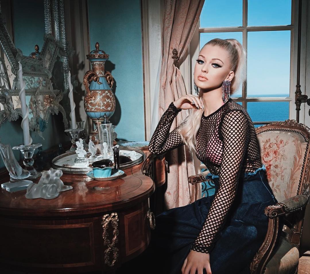 a young woman with blonde hair styled in a high ponytail is seated in a vintagestyle chair, posing indoors. she's wearing long, dangly earrings, a black net top with long sleeves, and a denim skirt. the interior is richly decorated with antique furniture, including a table adorned with a vase and various decorative items. a window offers a view of the sea in the background.