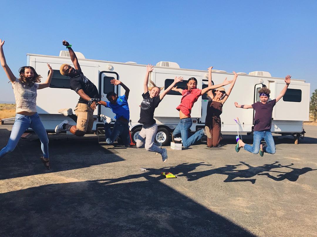 a group of eight teenagers are captured midair, jumping with exuberance in front of a white rv. their joy is palpable; arms flung wide, legs extended, they project a sense of carefree abandon. the bright sunlight and clear blue sky suggest a summer day, and the rv hints at a possible film set or outdoor event.  the teenagers' diverse appearances and clothing choices reflect a casual and heterogeneous group dynamic. their shadows stretch long on the ground behind them, adding depth to the image. the overall mood is lighthearted, playful, and celebratory.