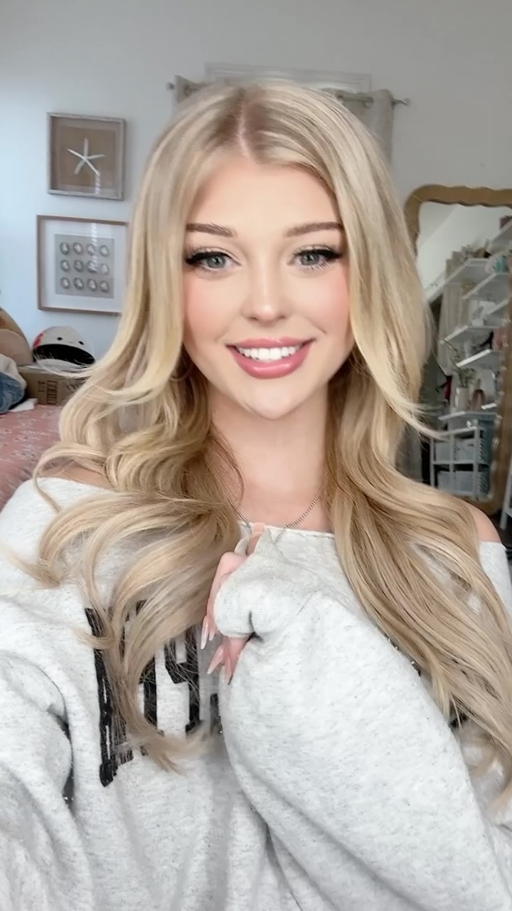 a portrait of loren gray, a young woman with long blonde hair, smiling and wearing a gray sweatshirt. she has makeup on, and her hair is styled with loose waves. she is posing indoors, and there is a mirror and other objects visible in the background.