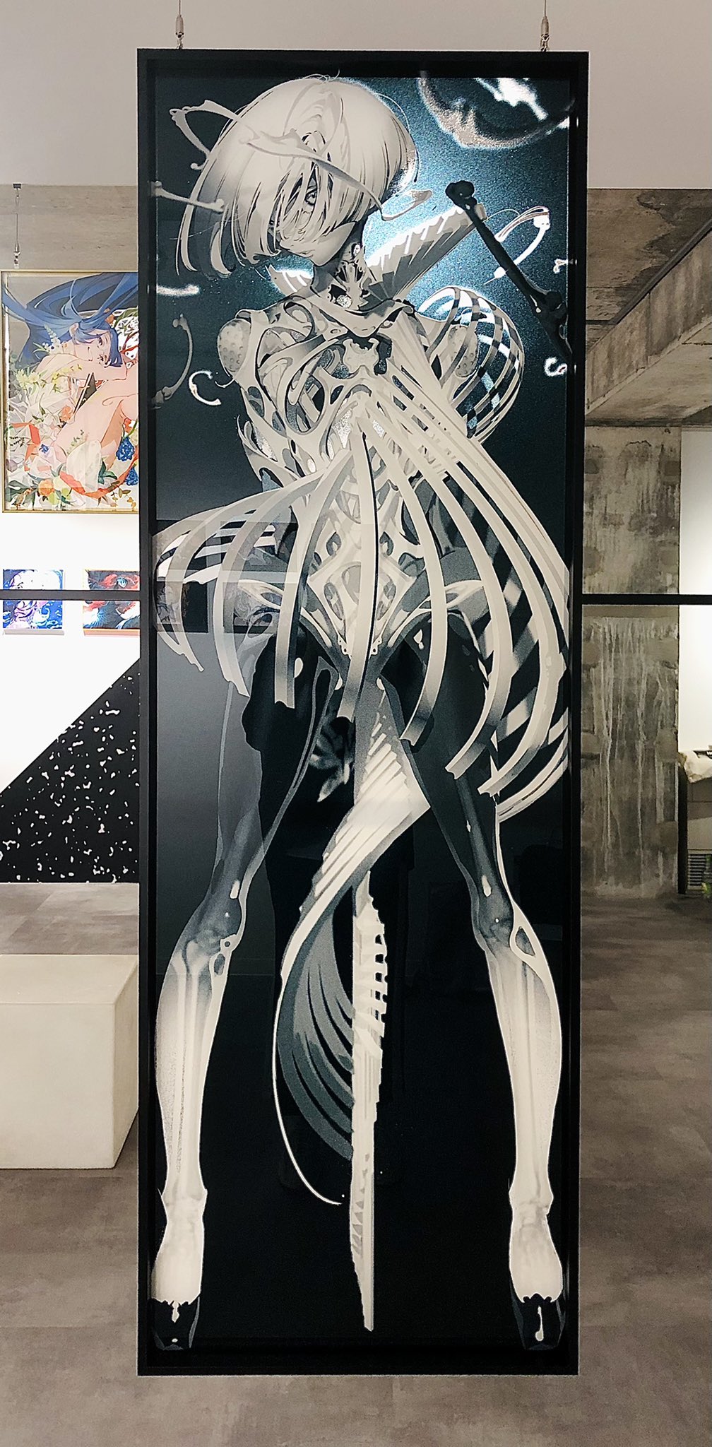 a large vertical artwork hangs in a gallery setting. the art depicts a stylized female figure in black and white, her body seemingly made of an intricate, skeletal structure reminiscent of both anatomy and futuristic machinery.  the background of the piece is dark, further highlighting the figure's stark white design. the gallery itself is minimalist, with gray floors and white walls featuring geometric black and white patterns. a second, smaller artwork with a more colorful anime style hangs on the wall in the background.