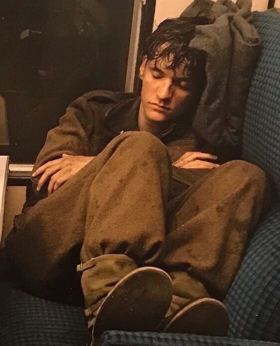 a young man in a worn military uniform is slumped in a train seat, sound asleep. his head is tilted back, a damp cloth draped over his face. his arms are wrapped around his legs, pulled up to his chest. the lighting is soft and warm, casting gentle shadows on his face. the overall palette evokes a sense of exhaustion and the somber atmosphere of wartime.