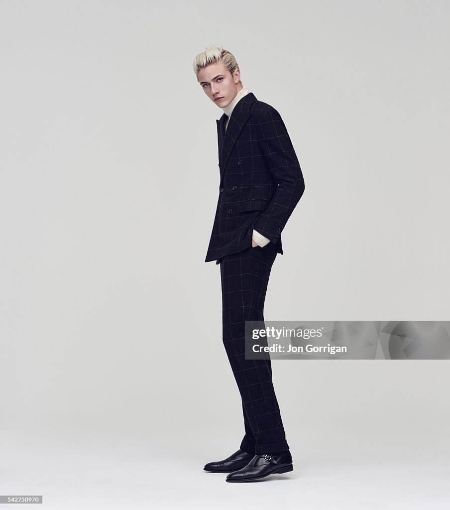 a full shot of a man in a suit. he is wearing a dark plaid suit with a white turtleneck underneath. he has blond hair and is looking at the camera. the man is standing in a studio.