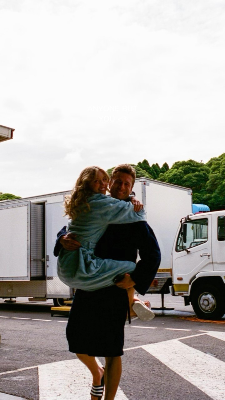 the image shows a man carrying a woman in his arms. they are both smiling happily and appear to be a couple. the woman is wearing a light blue robe and the man is wearing dark shorts and a dark robe. they are standing on a street with a crosswalk and some trucks parked nearby. the background includes trees and a cloudy sky. the overall setting suggests a film or movie set, given the presence of large trucks. the scene is warm and conveys a sense of love and affection.