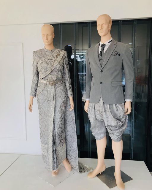 a bride and groom are standing in a traditional thai wedding attire. the bride is wearing a silver and gray dress with intricate details, while the groom is wearing a gray suit and silver and gray shorts. both are wearing  jewelry and standing on white mannequins. the couple is looking towards the left, the background has a blurry view of windows and white walls.