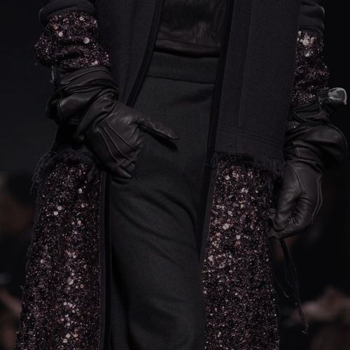 a closeup shot of a model wearing a black sequined outfit with black gloves. the model's outfit features a black sequined jacket with black leather gloves. the model is posing on a runway with a dramatic backdrop. the focus is on the model's hands and the detail of the sequined fabric. the image conveys a sense of style, elegance, and drama.