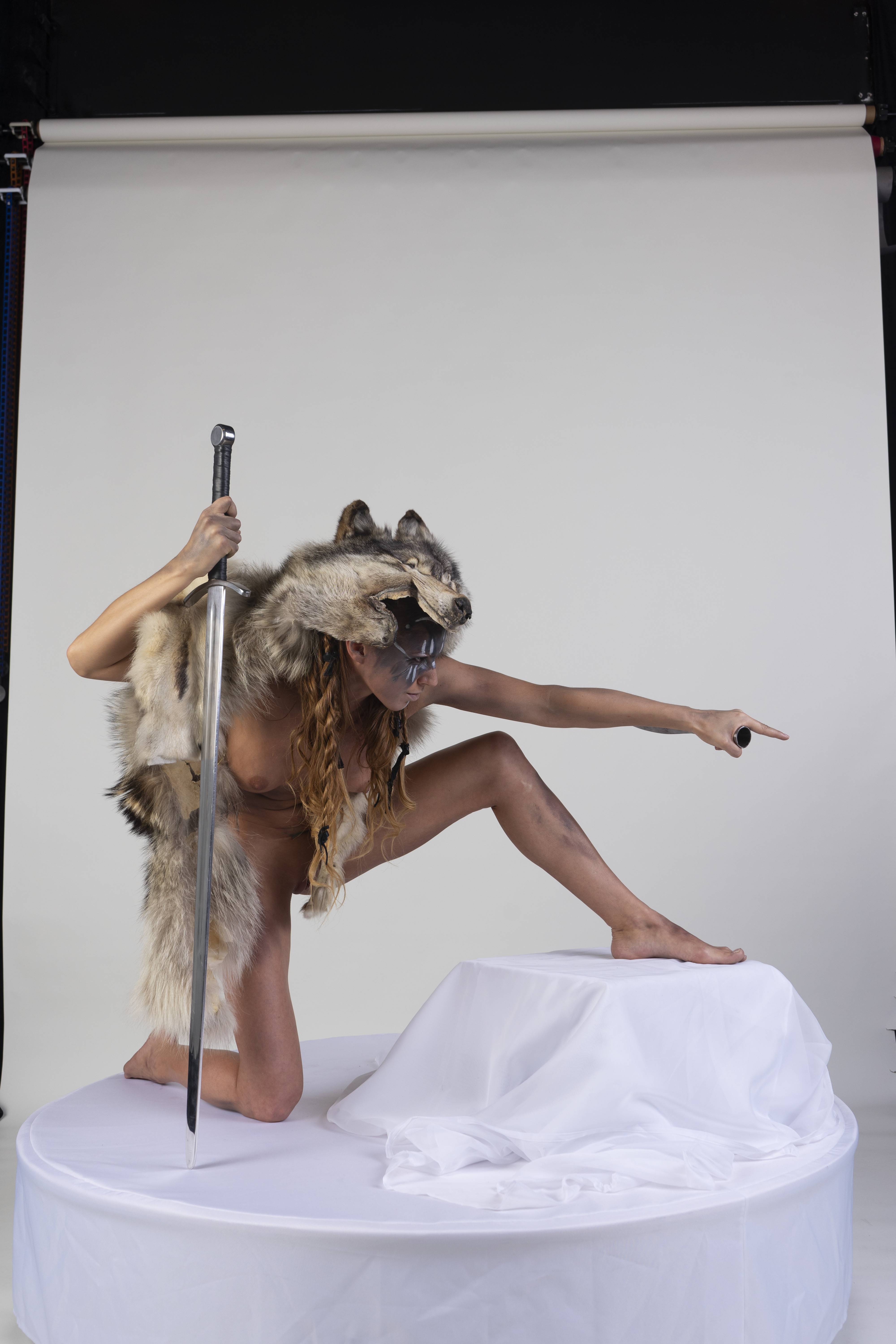 a woman dressed in a wolf pelt and loincloth is kneeling on a white platform in a studio. she is holding a sword in one hand and pointing with the other. she is wearing a wolf pelt headdress and has makeup on her face. the background is white.