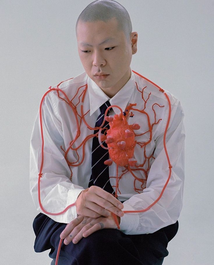 the photograph depicts a young asian man with a shaved head, seated and wearing a crisp white dress shirt and a dark tie.  a striking installation is the focal point: a realisticlooking red model of a human heart is affixed to his chest, connected by a network of red tubing that mimics the circulatory system.  the man's expression is serious and contemplative, his gaze directed downward. the background is a simple, unblemished white, emphasizing the subject and the artistic statement of the image. the overall aesthetic suggests a blend of fashion photography and conceptual art, exploring themes of the body, vulnerability, and perhaps internal states.