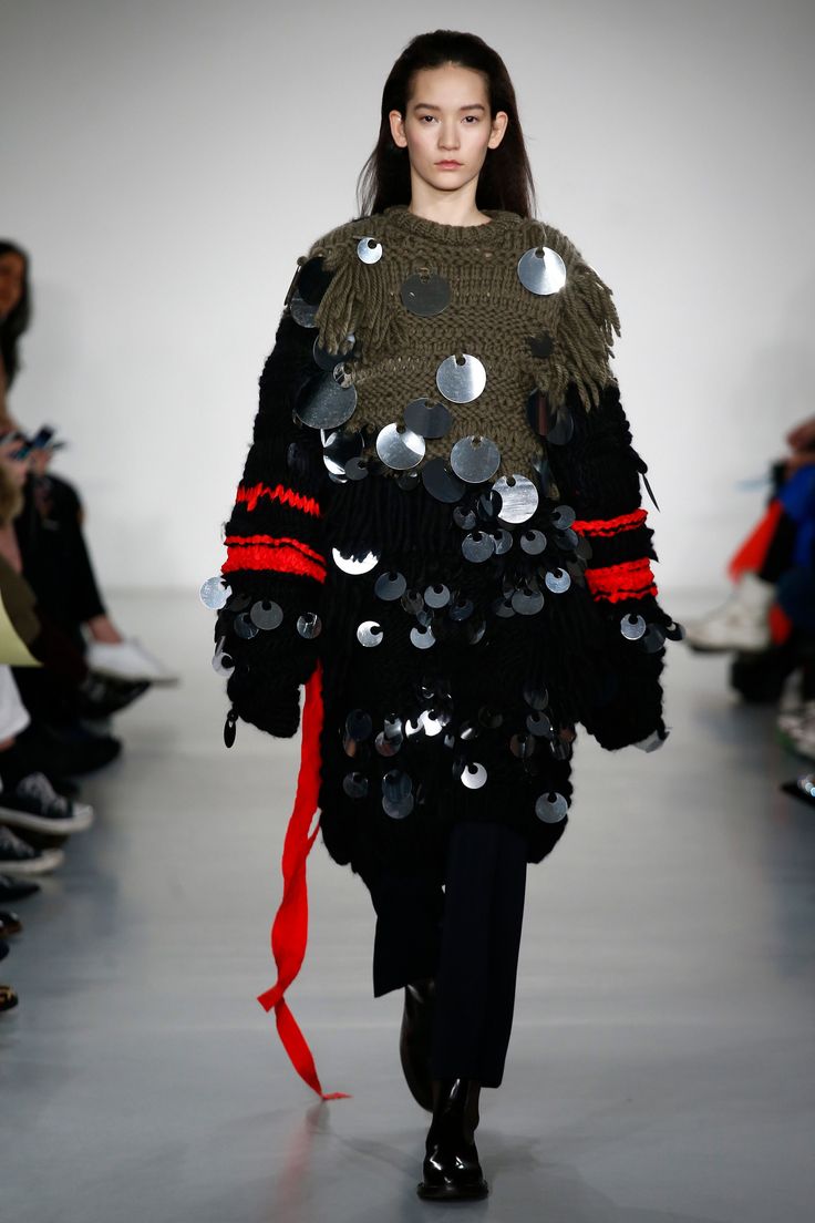 a fashion model walks the runway in a black and green sweater adorned with sequins, black pants, and black boots. the sweater is embellished with silver circles. the model has long black hair and a simple makeup look.
