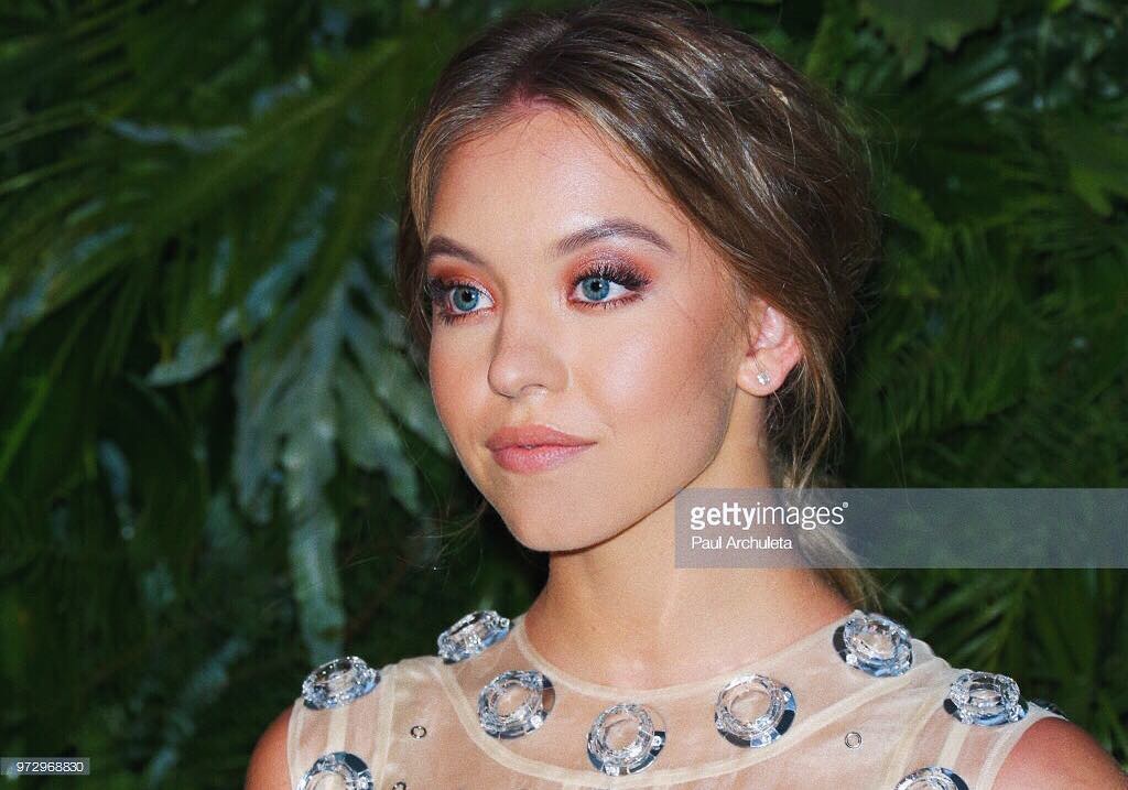 closeup portrait of a young woman with long, light brown hair styled in a loose low ponytail. she is wearing a beige dress adorned with crystal embellishments. her makeup is subtle and natural, emphasizing her blue eyes. the background is a blurry green foliage wall. the image is welllit and professionally taken, suggesting a promotional photoshoot or event photography.  the overall feeling is elegant and sophisticated.