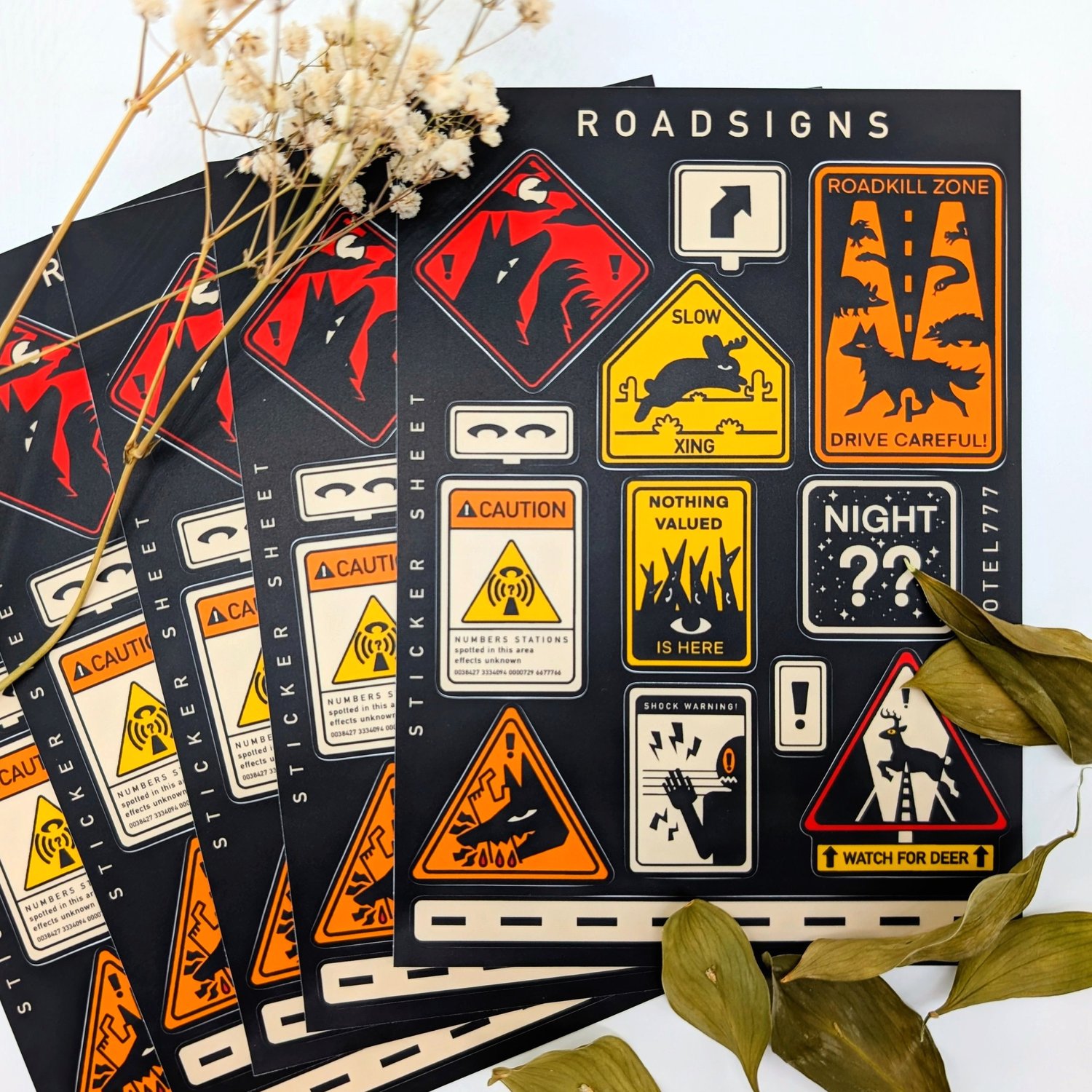 the image shows multiple sticker sheets with road sign designs. the sheets have a black background and feature a variety of quirky and illustrated road signs. some of the signs include warnings about "roadkill zone", "slow xing", "nothing valued is here", and "watch for deer". there are also signs with caution symbols and unusual illustrations. the overall aesthetic is a combination of warning signs and creative, graphic design. the sticker sheets are arranged in a stack and surrounded by dried flowers and leaves.