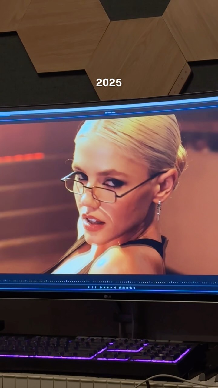 a blonde woman with glasses and earrings is displayed on a monitor. the number 2025 is superimposed on the image. a keyboard is visible below the monitor. the setting appears to be a studio.