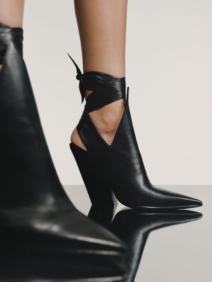 closeup image of a woman's legs showcasing a pair of black leather ankle boots. the boots are highheeled with a pointed toe and feature a unique cutout design at the ankle, secured with a leather tie wrapped around the leg. the boots appear to be made of highquality leather with a smooth finish. the reflection of the boots is visible on a glossy surface below.  the image emphasizes the stylish and elegant design of the footwear.