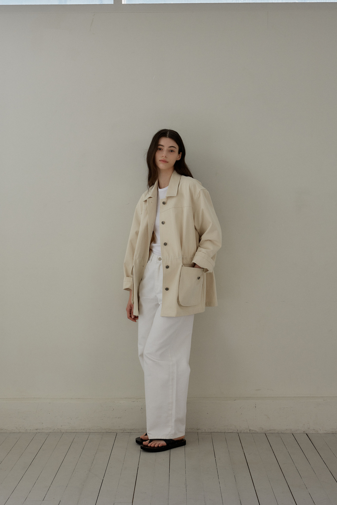a woman stands facing forward in a minimalist setting, wearing a casual outfit consisting of a lightcolored jacket, white pants, and black sandals. she has long, dark hair and a neutral expression. the background is a plain wall and wooden floor, emphasizing the simplicity and natural lighting of the scene.