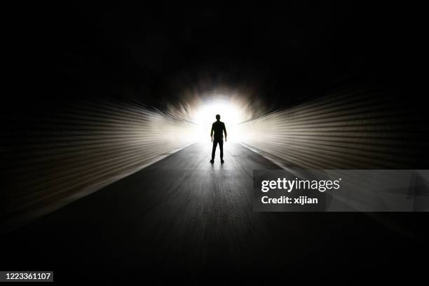 a silhouette of a person stands at the end of a tunnel with bright light, creating a strong contrast against the surrounding darkness. the pathway leading to the light source is visible in perspective.