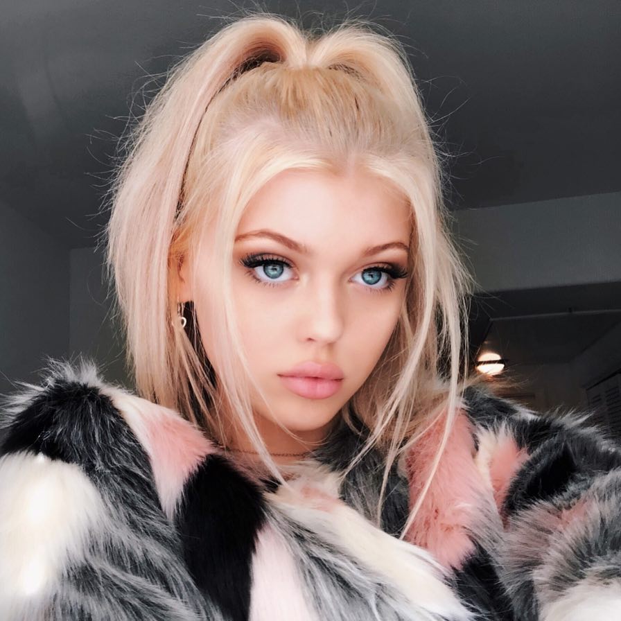 a portrait of a fairskinned woman with blonde hair styled in a halfup ponytail, striking blue eyes, and full makeup. she is wearing a multicolored fur coat with shades of pink, white, and black. she is also wearing heart shaped earrings. the background is slightly out of focus.
