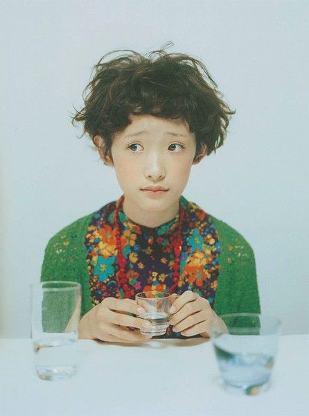 a young girl with short curly hair is sitting at a table, looking away from the camera. she is wearing a green sweater and a floral shirt and holding a glass of water in her hands. there are two more glasses of water on the table in front of her.  the image is taken from a slightly high angle, and the background is a light grey wall. the overall feeling of the photo is one of innocence and sweetness.