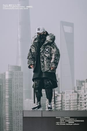 this is a fashion photo of a man wearing a camouflage winter jacket with fur on the hood. he is standing on the rooftop of a building in a city, showcasing his trendy street style. he is wearing a cap and black boots, and his stance and facial expression are confident and cool.