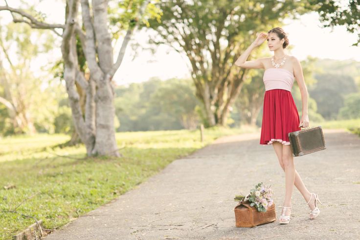 a young woman in a pink and red summer dress stands on a paved road, her hand shielding her eyes as she looks into the distance. she holds a vintage suitcase in her other hand, and a bouquet of flowers rests on a similar suitcase by her feet. lush green trees and grass line the road, creating a serene and picturesque setting. the sun bathes the scene in a warm, inviting glow, suggesting a sense of anticipation and the promise of adventure.