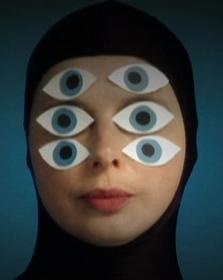 a closeup portrait of a woman wearing a black headscarf with multiple blue and white eyes pasted onto her face.