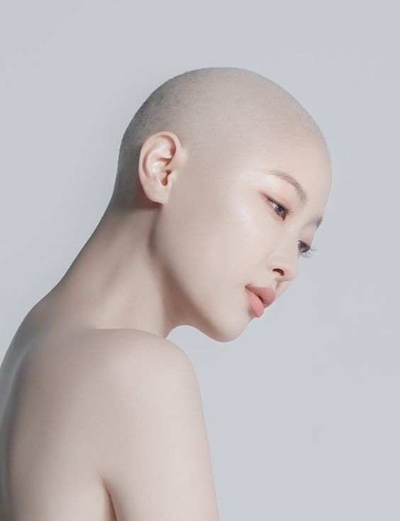 a closeup portrait of a woman with a shaved head, her head and shoulders are visible against a light grey background. the woman's gaze is directed downwards, creating a sense of serenity. the image has a minimalist aesthetic with soft lighting and a muted color palette, emphasizing the woman's natural beauty.