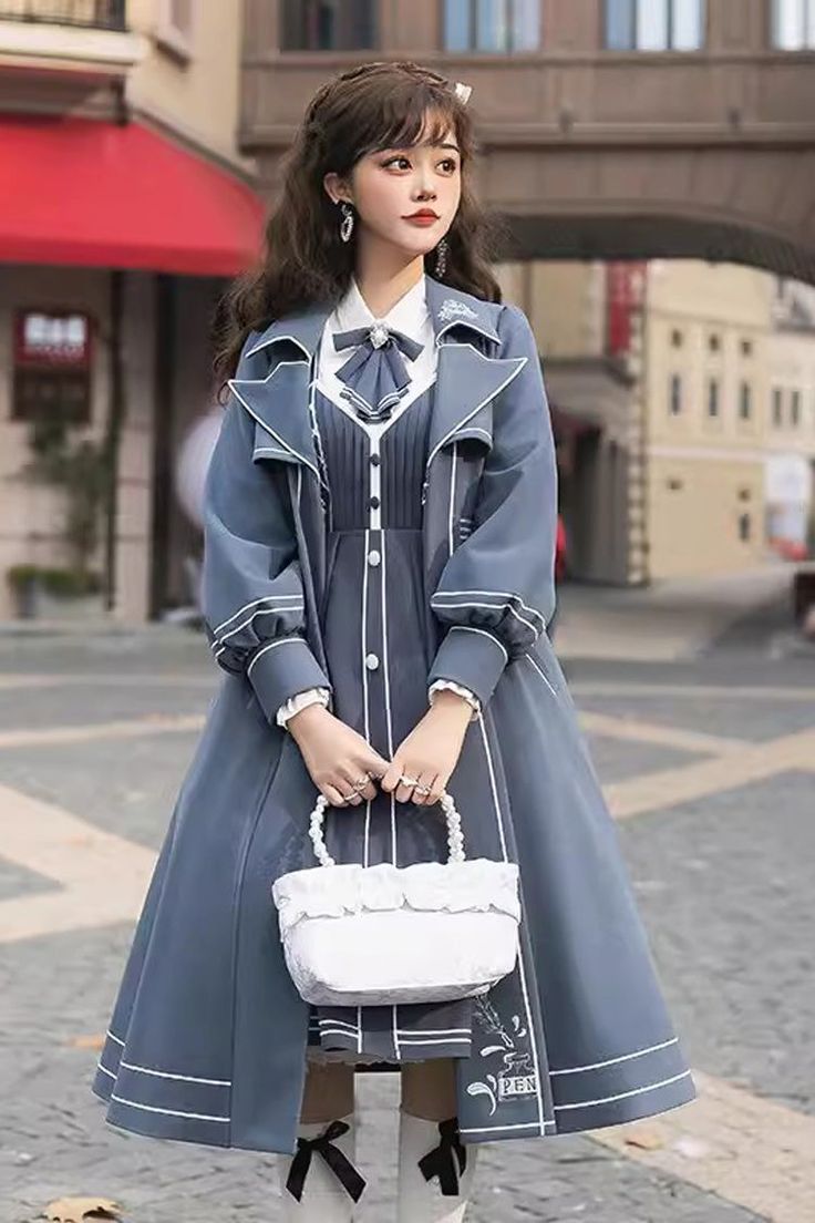 a young woman poses on a city street, showcasing a stylish outfit.  she wears a blue doublebreasted trench coat over a matching dress with white accents. the dress features a pleated bodice and a flowy skirt. she accessorizes with a white handbag, white socks, and black mary jane shoes. her dark hair is styled in loose waves, and she wears delicate earrings. the overall look is a charming blend of vintage and modern elements.