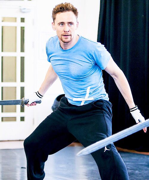 tom hiddleston, a wellknown actor, is seen in a training session, engaging in sword fighting. he's wearing a blue shirt and black pants, with white gloves on both hands. he's intensely focused on his practice, demonstrating his determination and skill in stage combat. the image captures the action and energy of the scene, showcasing the actor's dedication to his craft.