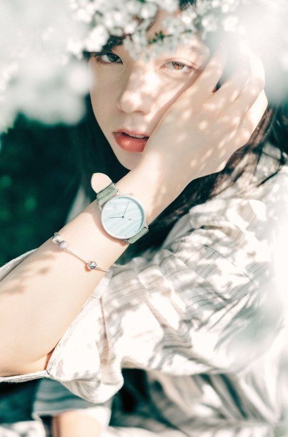 a woman is posing for a portrait, partially hidden behind white flowers. she is wearing a white shirt and a light blue watch. the image is lit by natural light, creating a soft and dreamy atmosphere.