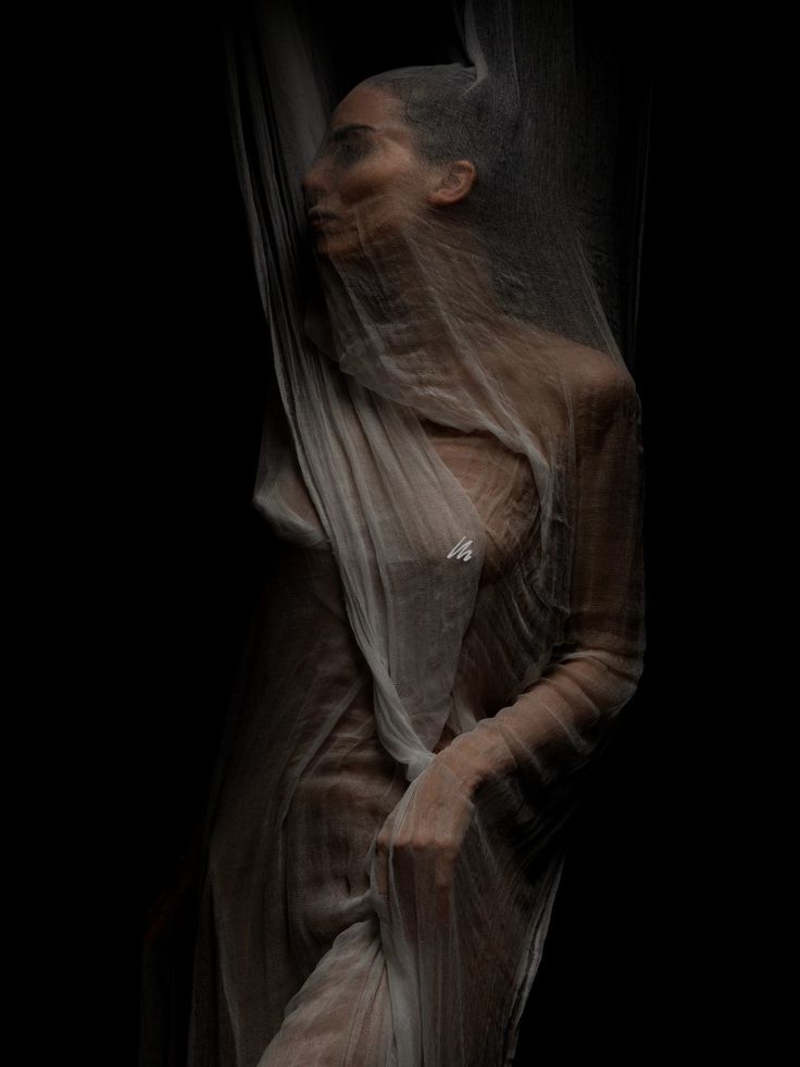 the image is a dark, moody portrait of a woman partially obscured by a sheer, draped fabric.  the fabric is lightcolored and loosely wraps around her body, obscuring much of her features, yet hinting at her form. the lighting is dramatic, with deep shadows and highlights creating a sense of mystery and intrigue. the overall aesthetic is artistic and evocative, suggestive of themes of concealment, vulnerability, and perhaps even a sense of ethereal beauty. the model's pose is contemplative, adding to the image's overall atmosphere.